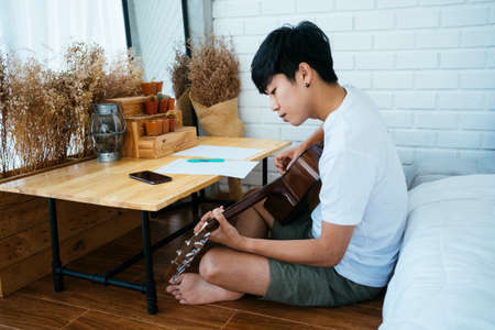 Black hair musician guy sitting at the wooden table in his bedroom and create his own song, he find the suitable note with guitar.の写真素材