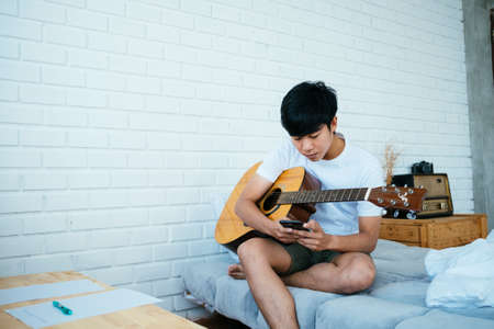 Black hair musician guy sitting on white bed in minimal bedroom and check his social media account while hug the guitar.の写真素材