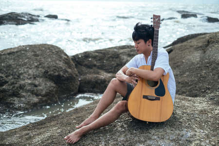 A musician man sitting on a rock looking at the sea with wooden guitar in his arm.