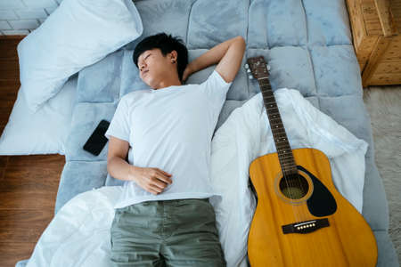 Black hair musician guy sleep on the white bed with guitar and smartphone.の写真素材