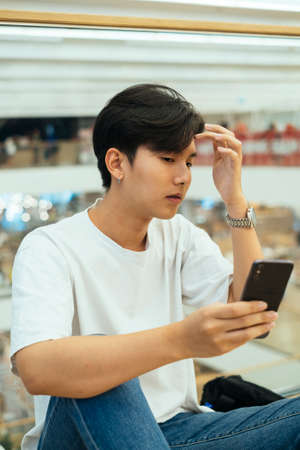 teenage guy in white t-shirt sitting in a mall holding his smartphone like an important device.