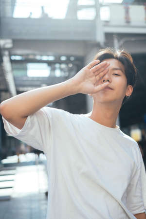 Teenage guy in white t-shirt standing in the sunlight raise his hand to hide his face.