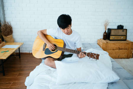 Black hair musician guy sitting on the white bed playing the guitar follow the note sheet.の写真素材