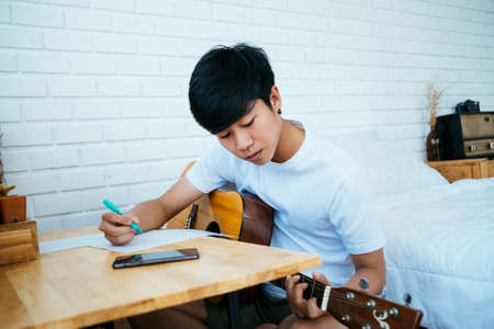 Black hair musician guy sitting at the wooden table in his bedroom and create his own song with guitar.の写真素材
