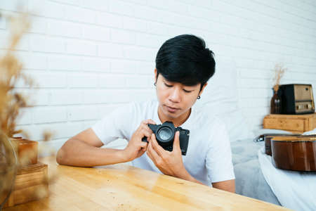 Black hair guy sitting at the wooden table looking at his camera in bedroom.の写真素材