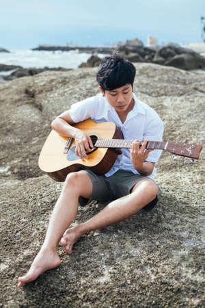 Black hair guy sitting on the rock at the sea and play the guitar.