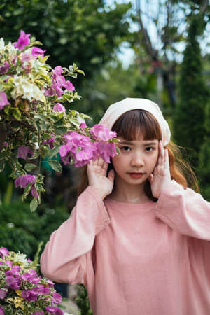 Brunette girl in beige headband and pink long sleeve t-shirt standing next to bougainvillea in the garden with trees.の写真素材