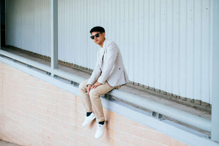 A man in white suit wearing sunglasses sitting on the handrail of the slope path of building.の写真素材
