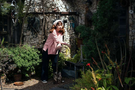 Brunette girl in beige headband and pink long sleeve t-shirt standing with spade in front of stone house in sunny day.の写真素材