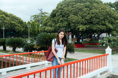 Long shot of casual woman in white t-shirt and jeans standing on the bridge with the garden as a...