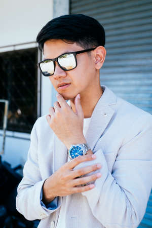 A man in white suit and sunglasses standing and touch his wrist and chin like a model.