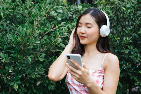 Beautiful young woman in headphones listening to music smiling with closed eyes at garden near green...