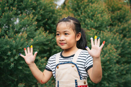 Beautiful girl playing with colors on fingers. Asian girl with hands painted in colorful paints ready for hand prints at garden.の写真素材