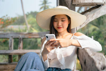 Cheerful asian woman on white shirt wearing hat and using smartphone with happy expression at countryside.の写真素材