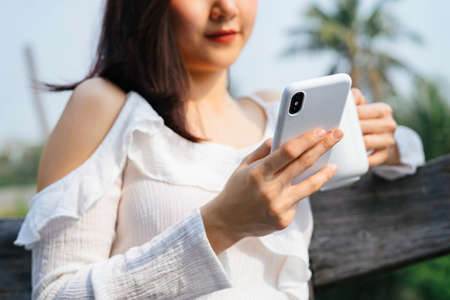 Close up asian woman on white shirt holding cup of coffee and using smartphone in nature.の写真素材