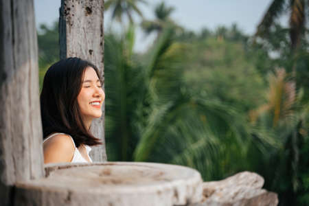 Cheerful asian woman on white shirt at wooden countryside hotel balcony. Beautiful nature on background.の写真素材