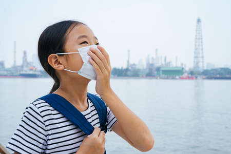 Sideview - Virus protection concept - asian little girl wearing protective mask, standing near river in smog day. Pollution every where. Covering her mouth with hand looking up the sky.の写真素材