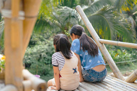 Back view - Two asian little girl sitting at the edge of wooden cafe made of bamboo tree, looking...