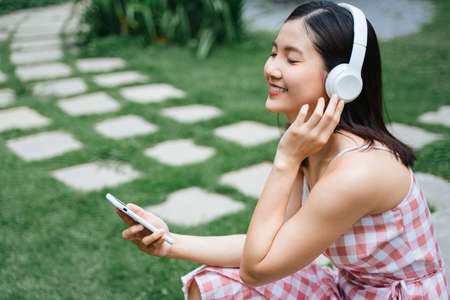 Beautiful young woman in headphones listening to music smiling with closed eyes sitting at garden near green tree plant.の写真素材