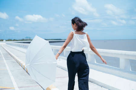 Portrait of young asian thai woman holding white umbrella, walking on the sea bridge in windy day.