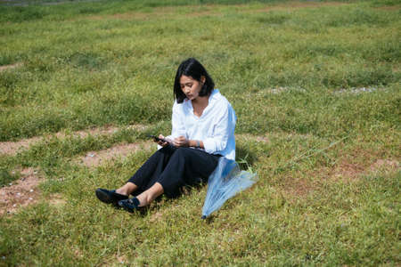 Cute young asian woman sitting on grasses field using smartphone with transparency umbrella beside...