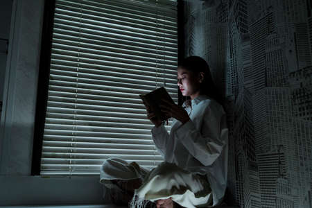Woman in white clothes sit at the window and read the book.