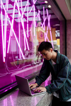 Short hair guy open his laptop to edit his work at the glass wall with pink neon light.