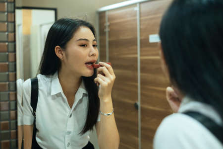 Black long hair college girl in uniform applying the lipstick on her lip in the restroom, close up.の写真素材