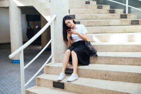 Black long hair college girl in uniform sitting on circle stair type her smartphone to text her friend.の写真素材