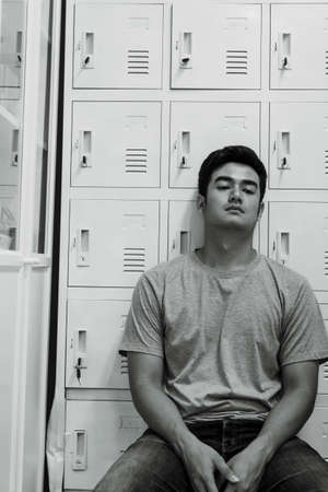 Black and White - Tired asian man on grey shirt and blue jeans sitting on a chair at locker room.
