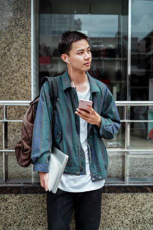 Short hair guy standing in front of the bulding holding smartphone while listening the song.