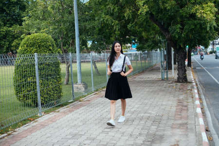 Black long hair college girl in uniform walking on the footpath at roadside near by the park.