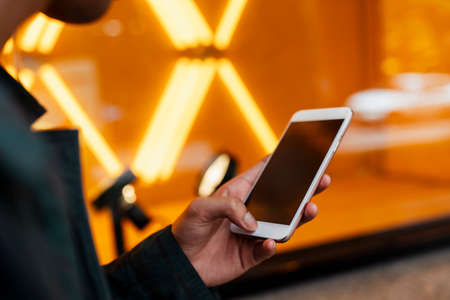 Cropped image of smartphone in a man's hands while he hold it in front of building with orange neon light.の写真素材