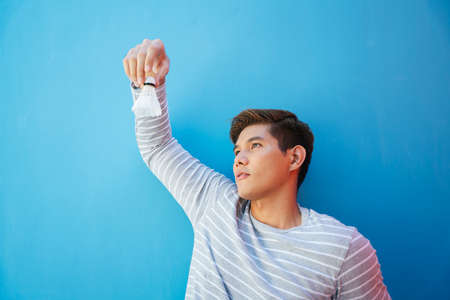 Young handsome asian guy holding shuttlecock with hand. Over blue background. Badminton player with...