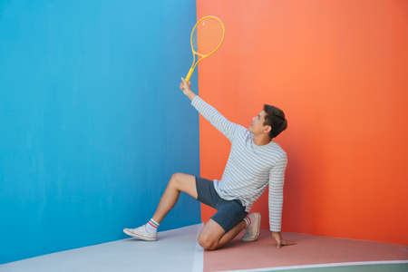 Attractive young asian man in casual costume holding tennis racket over blue orange background. Funny posing. Playing tennis. This is my racket gesture.の写真素材