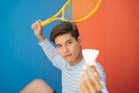 Attractive young asian man in casual costume holding tennis racket over blue orange background. Funny posing. Playing tennis. I am going to hit the shuttlecock gesture.の写真素材