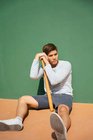 Asian baseball player with wooden bat sitting on the floor over green background.