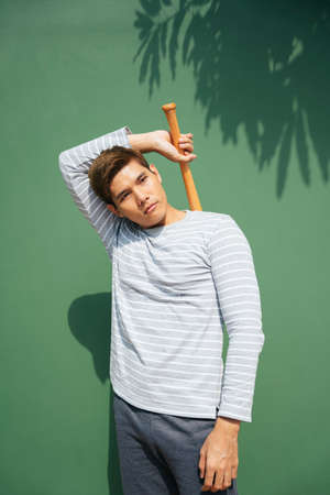 Young handsome asian man hold wooden baseball bat over green background. Baseball player on...