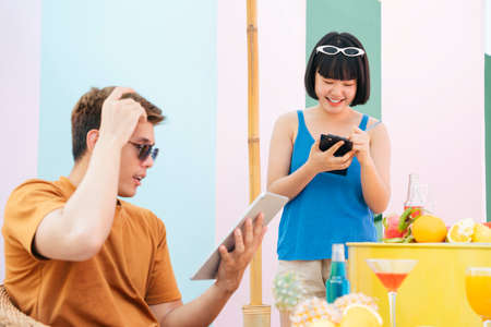 Young customer wearing sunglasses using tablet to order drink in tropical bar. Pretty waitress bob...