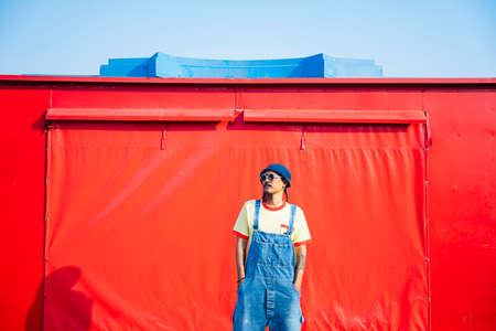 A man in yellow t-shirt and denim overalls standing and kick in front of the red trailer.の写真素材
