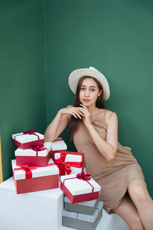 Sexy young asian woman wearing hat sitting with present gift boxes isolated over green background.