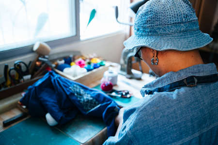 A man in blue clothes and hat, wearing apron sew the clothes for fixing.