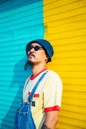 The man in yellow t-shirt and denim overalls standing and sitting in front of colorful shutter door in sunlight.の写真素材
