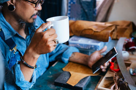 A man in blue shirt pause his leather work and drink the coffee to refresh himself, surfing on the internet.の写真素材