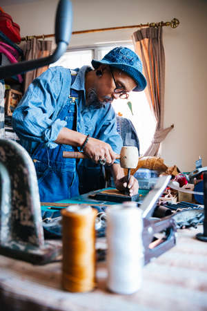A man in blue shirt and hat punching the leather before sewing.