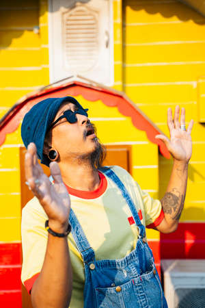 A man in yellow t-shirt and denim overalls standing in front of the to house in the sunlight with...