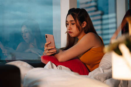 Beautiful mixed race woman using smartphone in bedroom near window.の写真素材