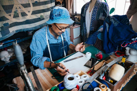 A man in blue clothes, hat and apron do the clothes cutting from the pattern on the paper.の写真素材