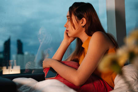 Beautiful mixed race woman sitting on a bed looking outside the window. Rainning in the city.の写真素材