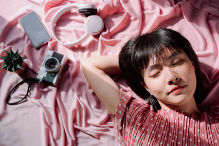 Top view - Beautiful asian thai woman short dark hair sleep over pink background. Camera, headphone, smartphone. Travel dreamer.の写真素材